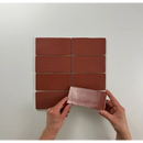 Rode-bruine Atlas Garnet Mate tegels in een grid met handen die een tegel plaatsen