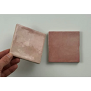 Een hand houdt een matte keramische tegel in zachte roze glans 10x10 met subtiele imperfecties trend tegels roze glans
